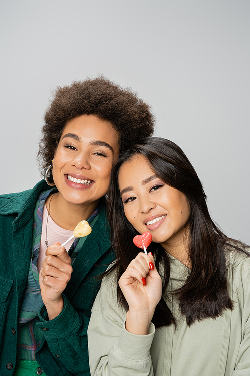 happy african american and asian women holding lollipops and looking at camera isolated on grey