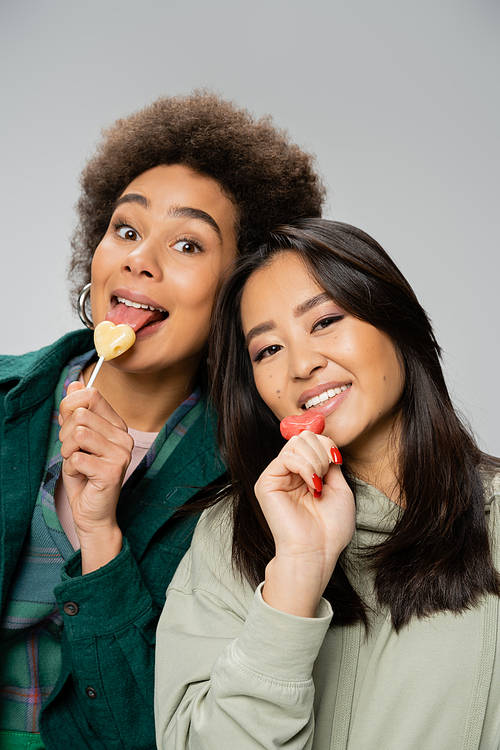cheerful african american woman with lollipop sticking out tongue near asian friend isolated on grey