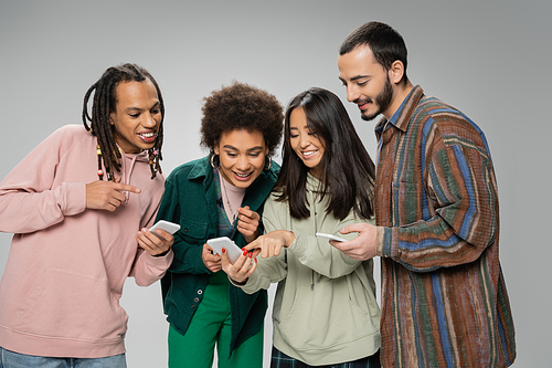 cheerful asian woman pointing at smartphone near trendy multiethnic friends isolated on grey