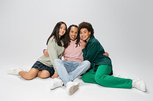 full length of trendy multiracial man hugging african american and asian women while sitting on grey background
