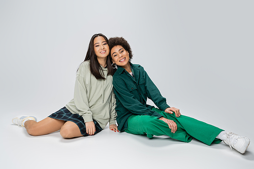 full length of asian and african american women in trendy outfit sitting and looking at camera on grey background