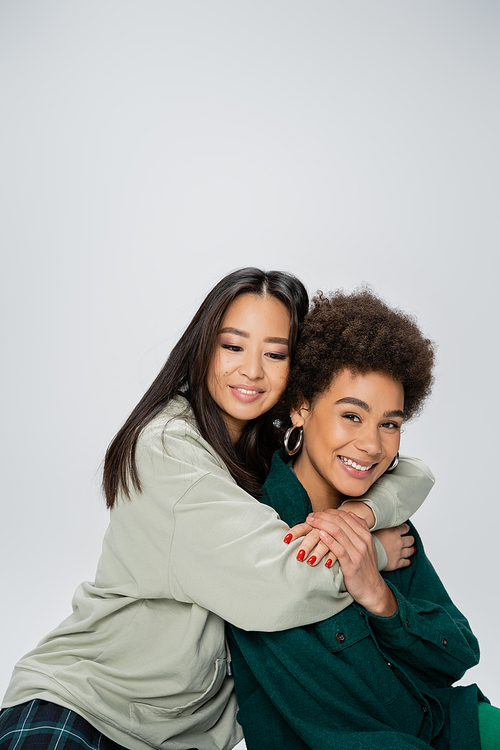 brunette asian woman hugging cheerful african american friend looking at camera isolated on grey
