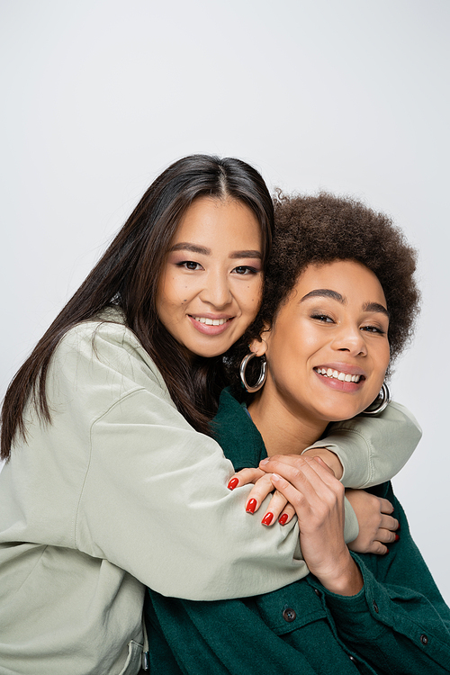 happy asian woman embracing curly african american friend while looking at camera isolated on grey