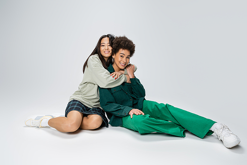 full length of happy asian woman embracing stylish african american friend while sitting on grey background