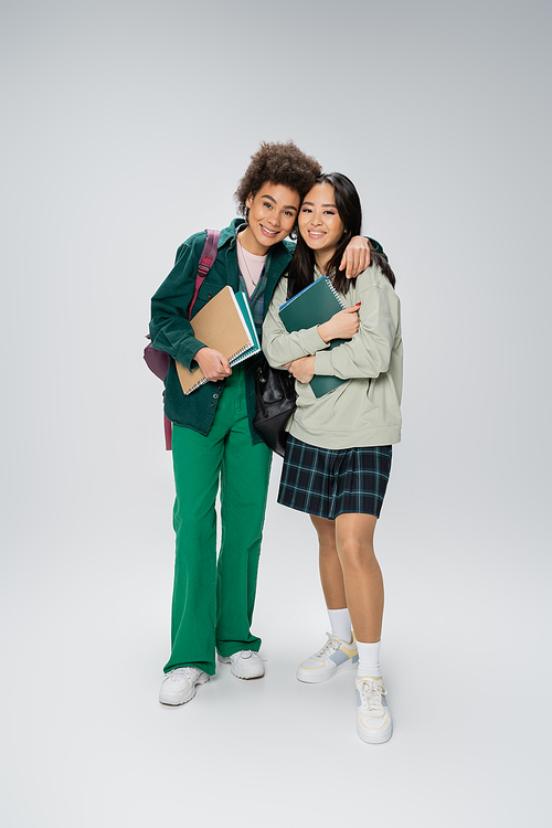 full length of african american and asian students with backpacks and copybooks smiling at camera on grey background