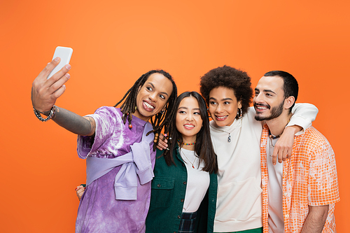 trendy man with dreadlocks taking selfie with happy multicultural friends isolated on orange