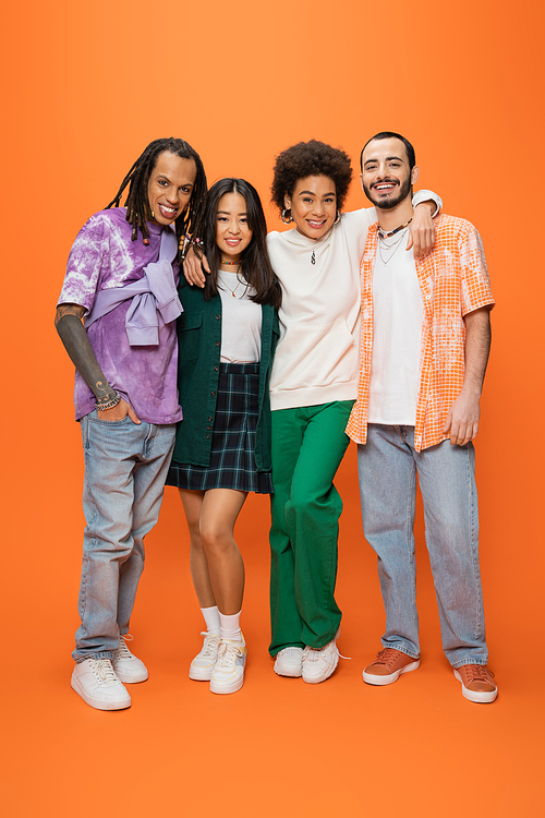 full length of fashionable multiethnic friends embracing and smiling at camera on orange background