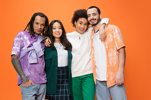 pleased african american woman hugging trendy multiethnic friends while looking at camera isolated on orange