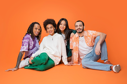 joyful multicultural friends in stylish clothes sitting and looking at camera on orange background