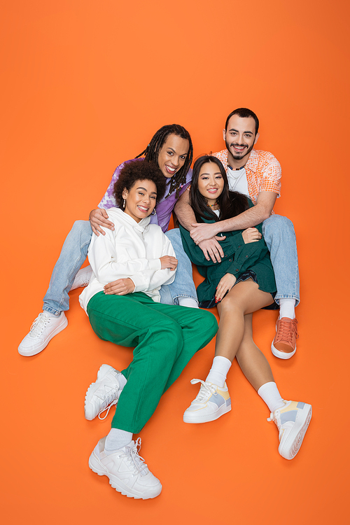 high angle view of happy and fashionable multiethnic friends sitting on orange background