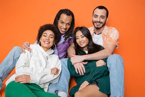 positive multicultural friends in trendy clothes smiling at camera while sitting on orange background