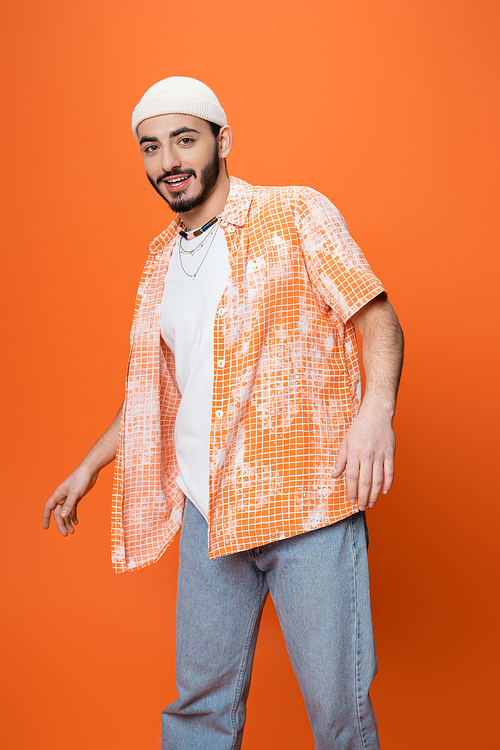 young bearded man in trendy casual attire smiling at camera while standing isolated on orange