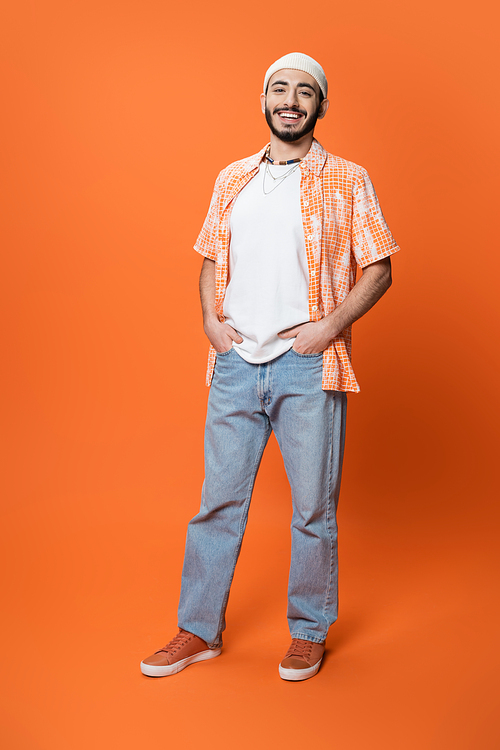 full length of happy bearded man in trendy shirt and white beanie posing with hands in pockets of blue jeans on orange background