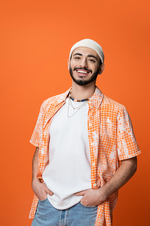 young bearded man in white beanie and beads posing with hands in pockets of jeans isolated on orange