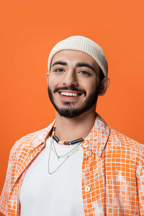 portrait of stylish bearded man in beanie and beads smiling at camera isolated on orange