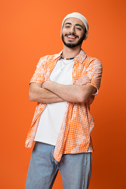 happy bearded man in trendy shirt and white beanie posing with crossed arms isolated on orange