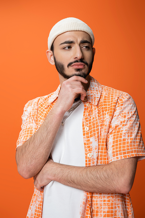 upset bearded man in stylish shirt and white beanie holding hand near chin and looking away isolated on orange
