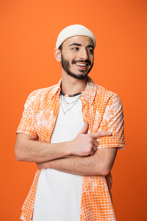 fashionable bearded man smiling and pointing with finger while looking away isolated on orange
