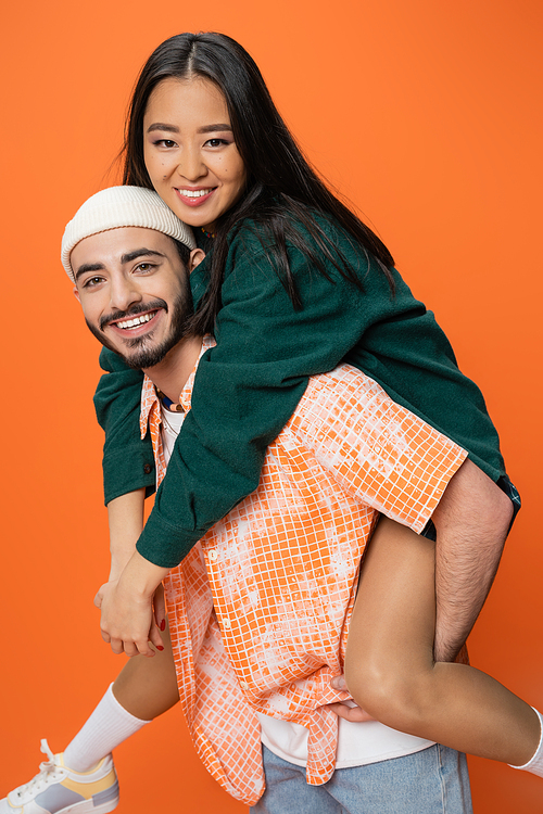 joyful bearded man in beanie piggybacking young asian woman while looking at camera isolated on orange