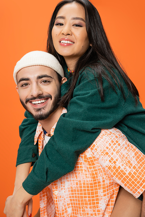 happy asian woman and young bearded man in beanie smiling at camera while having fun isolated on orange