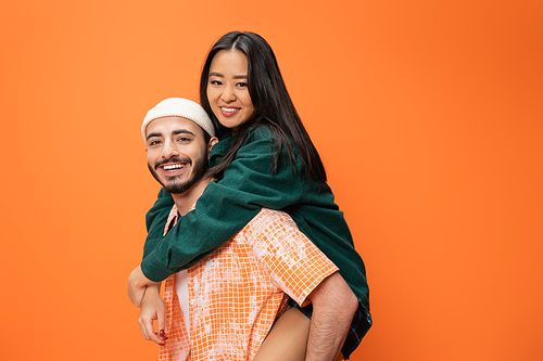 young bearded man in beanie piggybacking cheerful asian woman isolated on orange