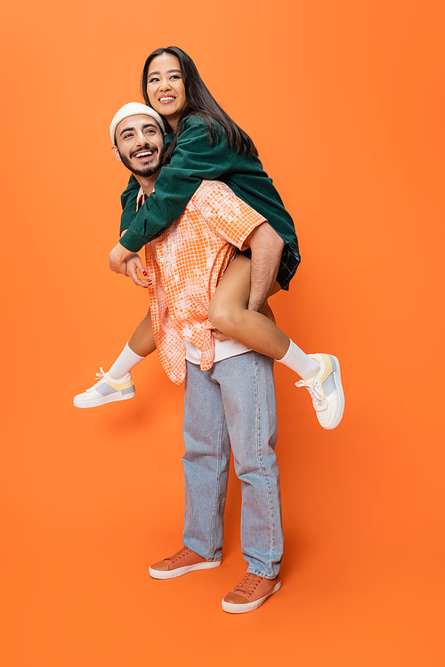 full length of excited man in beanie and jeans piggybacking trendy asian woman on orange background