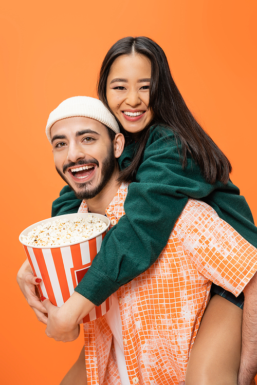cheerful man in beanie piggybacking asian woman holding bucket of popcorn isolated on orange