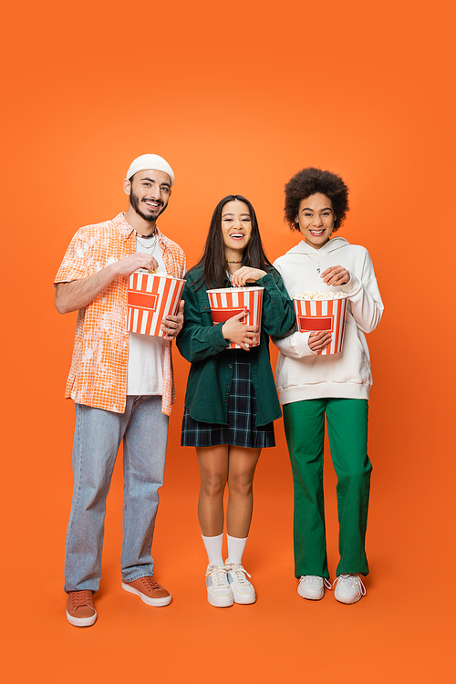 full length of trendy multiethnic friends standing with buckets of popcorn and smiling at camera on orange background