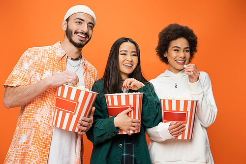 cheerful multiethnic friends in trendy clothes eating popcorn and looking away isolated on orange
