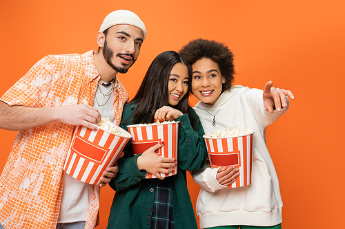 cheerful african american woman looking away and pointing with finger near multiethnic friends with popcorn isolated on orange