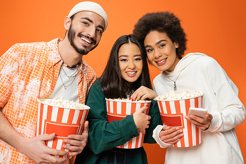 young and stylish multiethnic friends holding buckets of popcorn and smiling at camera isolated on orange