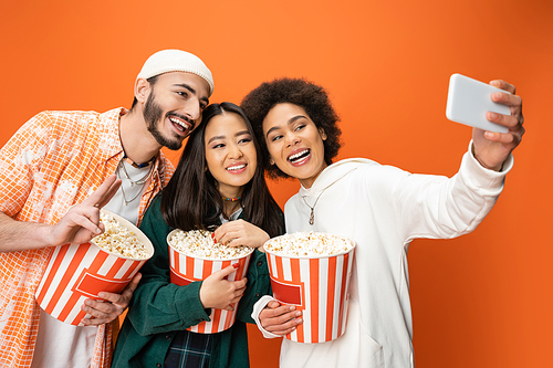 happy african american woman taking selfie with trendy friends holding buckets of popcorn isolated on orange