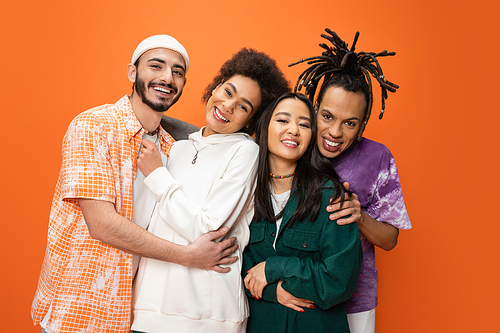 pleased multicultural friends in stylish outfit embracing and looking at camera isolated on orange