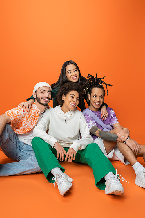 smiling asian woman hugging stylish multicultural friends sitting and looking away on orange background