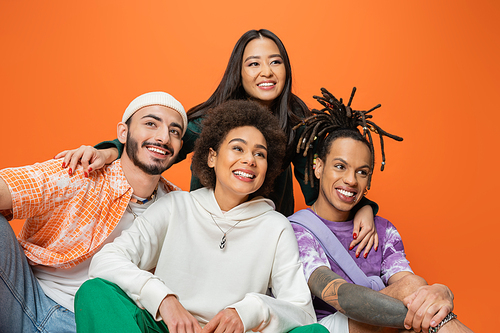 young asian woman embracing cheerful multicultural friends looking away isolated on orange