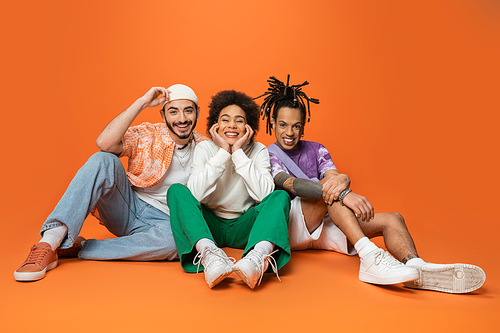 joyful multicultural friends in stylish outfit sitting and smiling at camera on orange background