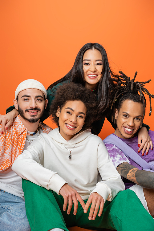 happy asian woman embracing stylish multiethnic friends smiling at camera isolated on orange