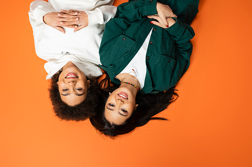 top view of happy african american and asian women in stylish attire lying on orange background