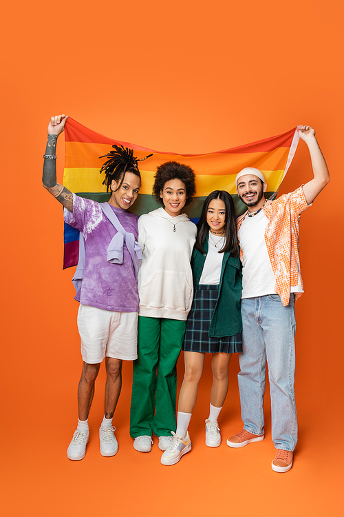full length of smiling multiethnic friends in trendy clothes holding lgbt flag on orange
