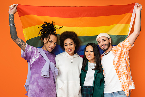 cheerful and trendy multiethnic friends posing with lgbt flag isolated on orange