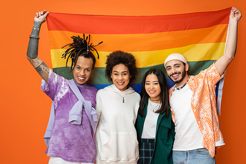happy and trendy multiracial friends looking at camera while standing with lgbt flag isolated on orange