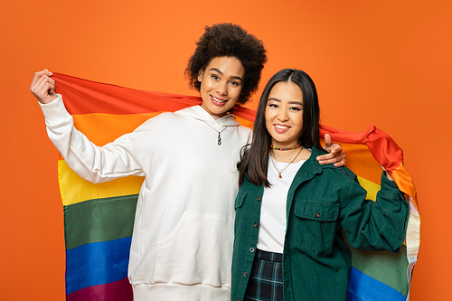 positive and trendy asian and african american women holding lgbt flag isolated on orange