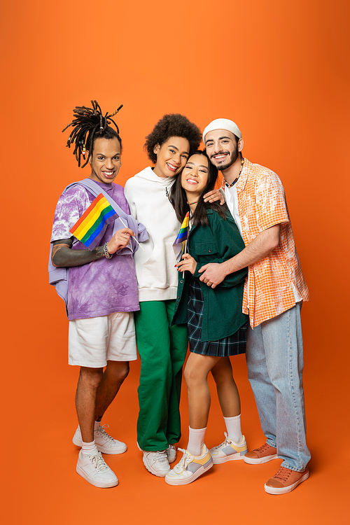 full length of stylish and cheerful multicultural friends embracing while holding lgbt flags on orange background