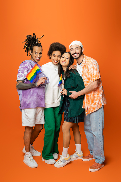 full length of multiracial friends in stylish outfit holding lgbt flags on orange background