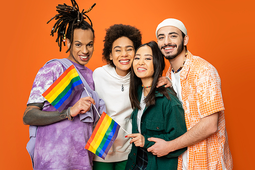 multicultural friends in stylish clothes holding lgbt flags and smiling at camera isolated on orange