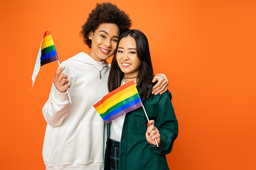 happy multiethnic women in trendy clothes holding lgbt flags isolated on orange