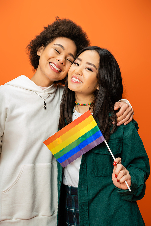 happy asian woman holding lgbt flag near african american friend smiling with closed eyes isolated on orange