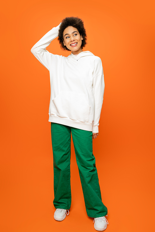 full length of happy african american woman in white hoodie and green pants looking away on orange background
