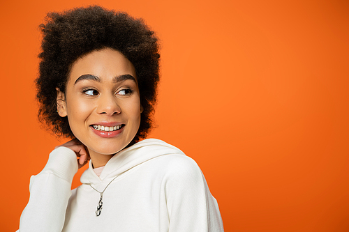 portrait of happy african american woman in white hoodie looking away isolated on orange