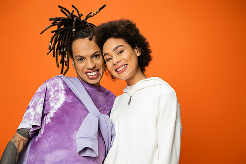 happy african american woman looking at camera near stylish friend with dreadlocks isolated on orange
